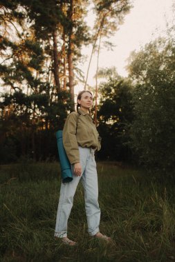 Doğayla bütünlük ve refah kavramı. Yoga antrenmanı için yoga minderiyle eğlenen aşırı neşeli kadın parkta meditasyon yapıyor. Kadın insan kavramı ve sağlıklı doğal yaşam tarzı. Dizinin bir parçası.