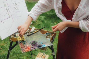 Bir kadının ellerini fırça ve boyalarla etekli bir tuvalin yanına yaklaştır. Farkındalık, sanat terapisi, yaratıcılık kavramı. Doğada plein hava boyama işlemi. Güzel sanatlar, eskiz, eskiz defteri..