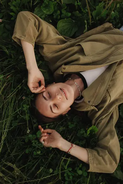 stock image Beautiful cute woman no makeup in white shirt laying down on green grass with white flowers. Wild nature, mood vacation, relax. Top view of girl. Be free, love forest. Part of the series.