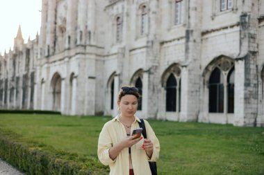 Middle aged traveling woman looks for the way using the online map on her phone and passes the Monteiro dos Jeronimo's in Belem, Lisbon, Portugal. Travel and solo tourism concept. Part of the series. clipart