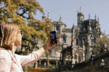 Gözlem güvertesindeki orta yaşlı gezgin kadın Portekiz 'deki Quinta da Regaleira' nın güzel bir görüntüsünü cep telefonuyla çekiyor. Seyahat ve solo turizm konsepti. Dizinin bir parçası.