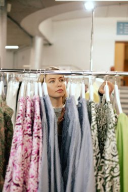 Middle aged woman chooses a dress made of natural linen in a shopping mall. Close-up of lady plucked hanger choosing clothes in clothing store. Sale promotion and shopping concept. Part of the series. clipart