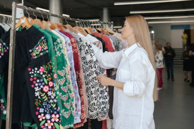 Orta yaşlı bir kadın alışveriş merkezinde iplikli bir gömlek rafının üzerinde koşuyor. Ukrayna ulusal kıyafetleri. Elbise mağazasından elbise seçen kadın ellerinin yakın çekimi.