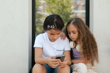 Çok ırklı ortaokul. Sırt çantalı kızlar okul bahçesinde telefon ve kulaklık kullanıyor. Alfa Geni. Bir serinin parçası.