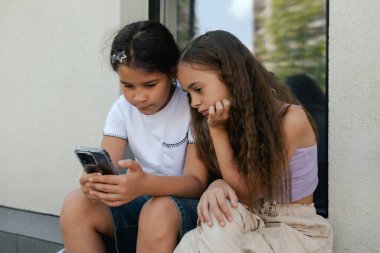 Çok ırklı ortaokul. Sırt çantalı kızlar okul bahçesinde telefon ve kulaklık kullanıyor. Alfa Geni. Bir serinin parçası.