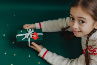 Küçük tatlı bir kız elinde kağıt el işi hediyesiyle arka planda Noel süsleri tutuyor. Alfa Geni. Bir serinin parçası.