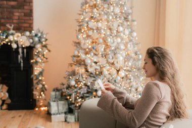 Çekici bir kadının Noel tebrik kartı okuduğu yan manzara. Arka planda, rahat oturma odasında, şenlikli bir iç mekanı olan Xmas ağacı. Evde mektup okumaktan hoşlanan bir bayan şaşırdım. Bir serinin parçası.