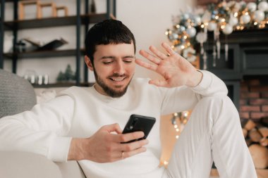 Mutlu adam evde akıllı telefon kullanarak sanal Noel videosunu kutluyor. Ailesini ve arkadaşını arıyor. Çevrimiçi sanal tatil buluşması, normal ve sosyal uzaklık. Bir serinin parçası.