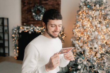 Sakallı bir adamın, Noel tebrik kartı okurken rahat oturma odasında, şenlikli bir şekilde oturduğu Noel ağacının arka planında görüntüsü. Evde mektup okumaktan hoşlanan bir bayan şaşırdım. Bir serinin parçası.