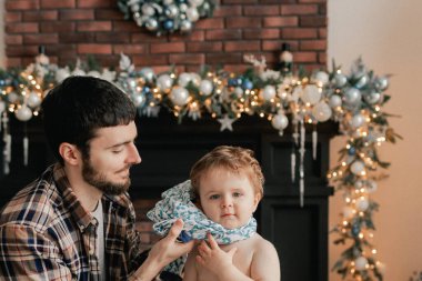 Genç bir baba, oğlunun oturma odasındaki Noel ağacının yanında kazağını değiştirmesine yardım eder. Yalnız ebeveynlik kavramı ve yeni yıl için hediyeler, anneler günü ve babalar günü. Modern iç mekan. Bir serinin parçası.