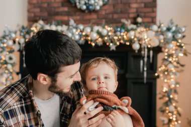 Genç bir baba, oğlunun oturma odasındaki Noel ağacının yanında kazağını değiştirmesine yardım eder. Yalnız ebeveynlik kavramı ve yeni yıl için hediyeler, anneler günü ve babalar günü. Modern iç mekan. Bir serinin parçası.