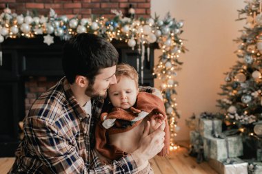 Genç bir baba, oğlunun oturma odasındaki Noel ağacının yanında kazağını değiştirmesine yardım eder. Yalnız ebeveynlik kavramı ve yeni yıl için hediyeler, anneler günü ve babalar günü. Modern iç mekan. Bir serinin parçası.