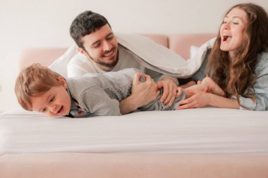 Mutlu bir aile yatakta birlikte gülüyor ve eğleniyor. İki ebeveyn evde oğluyla oynuyor. Anne ve baba hafta sonları çocuklarıyla kaliteli zaman geçiriyorlar. Bir serinin parçası.