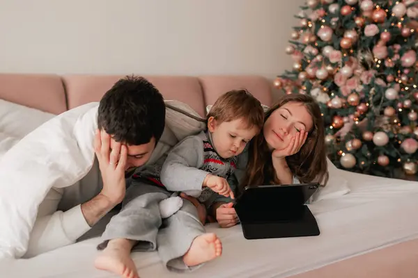 Tired parents with their son playing on a tablet on the bed. Two parents playing with their son in tablet at home. Part of a series.