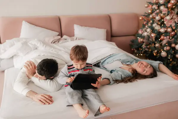 Tired parents with their son playing on a tablet on the bed. Two parents playing with their son in tablet at home. Part of a series.