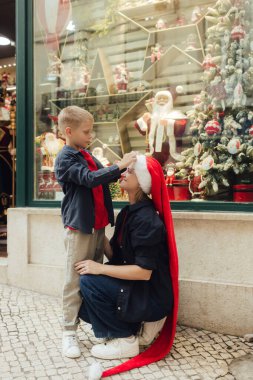 Noel pazarında annesi olan tatlı bir okul çocuğu. Moda kıyafetleri içindeki neşeli çocuk vitrinlere hediyelerle süslenmiş şeyler yapıyor. Tatiller, Noel, çocukluk ve insanlar konsepti.