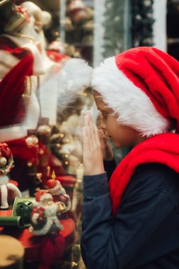 Noel Baba şapkalı küçük okul çocuğu. Hediyelik eşya dükkanının vitrinine bakar ve ailesi için sürprizleri seçer. Tatiller, Noel, çocukluk ve insanlar konsepti.