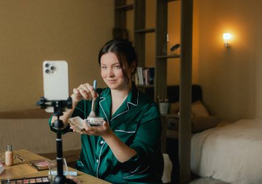 Artı, sosyal medya için akıllı telefondan vlog kaydederken yüzüne fırçayla makyaj yapan boyda bir kadın. Çok ırklı makyaj sanatçısı vlogger canlı yayın makyaj eğitmeni. Dizinin bir parçası. UGC.