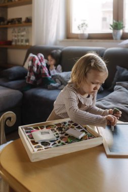 Oyun odasında iki kız kardeş. Genç bir kız tabletle oynuyor ve küçük bir kız yaratıcı oyun zamanının tadını çıkarıyor. Evinde rahat ve aydınlık bir ortamda. Jenerasyon alfa kavramı. Bir serinin parçası.