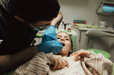 Saçları örgülü, dişçi koltuğunda oturmuş kameraya bakan küçük bir kızın kesilmiş portresi. Eldivenli bir doktor muayene aletleri bulundurur. Çocuk dişçiliği. Bir serinin parçası.