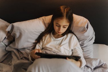 Güzel genç kız tablet üzerinde uygulama kullanıyor, çevrimiçi video görüşmesinde konuşuyor, gülüyor, gizlice film izliyor, online oyunlar oynuyor, yatak örtülerinin altında yatıyor. Jenerasyon alfa kavramı. Bir serinin parçası