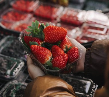 Kadın alıcıların elinde taze böğürtlenli şeffaf plastik kaplar. Yerel pazarda paketler üst üste istiflenir. Çilekler sert, zengin ve parlaktır..