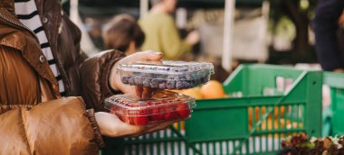 Kadın alıcıların elinde taze böğürtlenli şeffaf plastik kaplar. Yerel pazarda paketler üst üste istiflenir. Çilekler sert, zengin ve parlaktır..