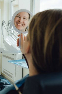 Dişçi koltuğunda ayna ile gülümseyen olgun bir kadın. Klinikte randevu. Diş beyazlatma, temizlik ya da oral, hastanede ağız ve sakız bakımı. Yumuşak odaklanma. Dizinin bir parçası. Yakın plan..