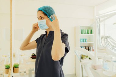 Randevuya hazırlanan genç dişçi maske takıyor. Tıpta kadın, diş bakımı, klinikte check-up. Diş beyazlatma, temizlik ya da oral, ağız ve sakız bakımı. Bir serinin parçası.