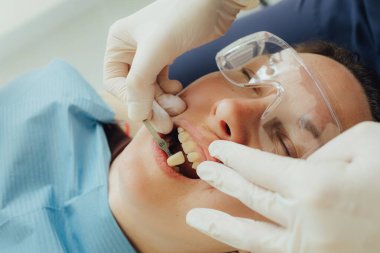 Genç bir dişçiye dişçi muayenesinde gülümseyen olgun bir kadın. Klinikte randevu. Diş beyazlatma, temizlik ya da oral, hastanede ağız ve sakız bakımı. Yumuşak odaklanma. Dizinin bir parçası. Yakın plan..