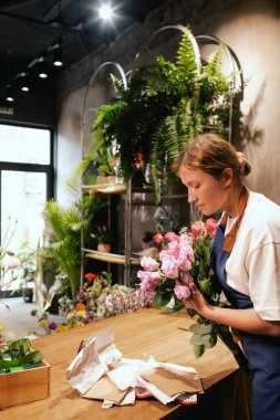 Önlüklü orta yaşlı kadın çiçekçi çiçekçide çiçek buketi için çiçek düzenliyor. Küçük bir yerel işletme. Dizinin bir parçası. Yumuşak odak