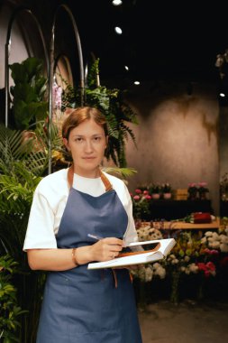 Önlüklü orta yaşlı kadın çiçekçi akıllı telefondan arıyor. Lojistiği ve teslimatı ayarlıyor, emirleri alıyor. Küçük bir yerel işletme. Dizinin bir parçası. Yumuşak odak