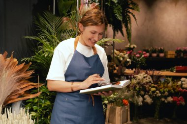 Önlüklü orta yaşlı kadın çiçekçi akıllı telefondan arıyor. Lojistiği ve teslimatı ayarlıyor, emirleri alıyor. Küçük bir yerel işletme. Dizinin bir parçası. Yumuşak odak