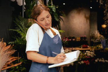 Önlüklü orta yaşlı kadın çiçekçi akıllı telefondan arıyor. Lojistiği ve teslimatı ayarlıyor, emirleri alıyor. Küçük bir yerel işletme. Dizinin bir parçası. Yumuşak odak
