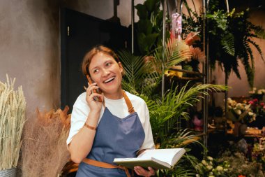 Önlüklü orta yaşlı kadın çiçekçi akıllı telefondan arıyor. Lojistiği ve teslimatı ayarlıyor, emirleri alıyor. Küçük bir yerel işletme. Dizinin bir parçası. Yumuşak odak