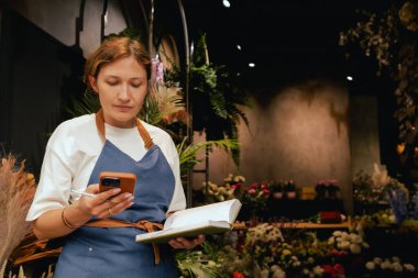 Önlüklü orta yaşlı kadın çiçekçi akıllı telefondan arıyor. Lojistiği ve teslimatı ayarlıyor, emirleri alıyor. Küçük bir yerel işletme. Dizinin bir parçası. Yumuşak odak