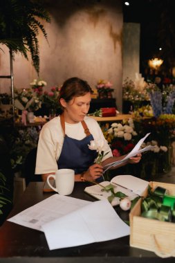 Orta yaşlı bir kadın, arka planda çeşitli bitkiler bulunan bir çiçekçide hesap makinesinin mali durumunu hesaplar. Lojistiği ve teslimatı ayarlıyor, emirleri alıyor. Küçük yerel işletme