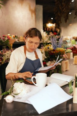 Orta yaşlı bir kadın, arka planda çeşitli bitkiler bulunan bir çiçekçide hesap makinesinin mali durumunu hesaplar. Lojistiği ve teslimatı ayarlıyor, emirleri alıyor. Küçük yerel işletme