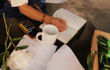 Orta yaşlı bir kadın, arka planda çeşitli bitkiler bulunan bir çiçekçide hesap makinesinin mali durumunu hesaplar. Lojistiği ve teslimatı ayarlıyor, emirleri alıyor. Küçük yerel işletme