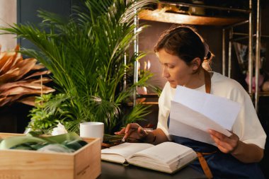 Orta yaşlı bir kadın, arka planda çeşitli bitkiler bulunan bir çiçekçide hesap makinesinin mali durumunu hesaplar. Lojistiği ve teslimatı ayarlıyor, emirleri alıyor. Küçük yerel işletme