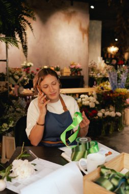 Önlüklü orta yaşlı kadın çiçekçi akıllı telefondan arıyor. Lojistiği ve teslimatı ayarlıyor, emirleri alıyor. Küçük bir yerel işletme. Dizinin bir parçası. Yumuşak odak