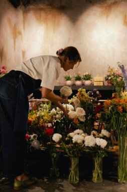 Önlüklü orta yaşlı kadın çiçekçi çiçekçide çiçek buketi için çiçek düzenliyor. Küçük bir yerel işletme. Dizinin bir parçası. Yumuşak odak
