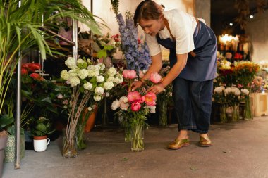 Önlüklü orta yaşlı kadın çiçekçi çiçekçide çiçek buketi için çiçek düzenliyor. Küçük bir yerel işletme. Dizinin bir parçası. Yumuşak odak
