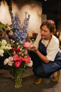 Önlüklü orta yaşlı kadın çiçekçi çiçekçide çiçek buketi için çiçek düzenliyor. Küçük bir yerel işletme. Dizinin bir parçası. Yumuşak odak