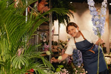 Önlüklü orta yaşlı kadın çiçekçi çiçekçide çiçek buketi için çiçek düzenliyor. Küçük bir yerel işletme. Dizinin bir parçası. Yumuşak odak