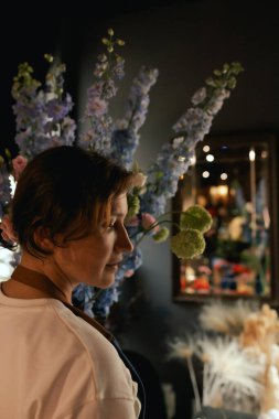 Önlüklü orta yaşlı kadın çiçekçi çiçekçide çiçek buketi için çiçek düzenliyor. Küçük bir yerel işletme. Dizinin bir parçası. Yumuşak odak