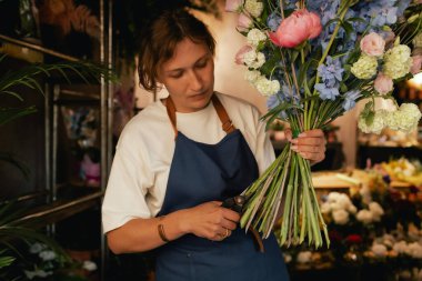 Önlüklü orta yaşlı kadın çiçekçi çiçekçide çiçek buketi için çiçek düzenliyor. Küçük bir yerel işletme. Dizinin bir parçası. Yumuşak odak