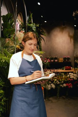 Önlüklü orta yaşlı kadın çiçekçi akıllı telefondan arıyor. Lojistiği ve teslimatı ayarlıyor, emirleri alıyor. Küçük bir yerel işletme. Dizinin bir parçası. Yumuşak odak