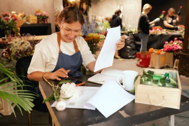 Orta yaşlı bir kadın, arka planda çeşitli bitkiler bulunan bir çiçekçide hesap makinesinin mali durumunu hesaplar. Lojistiği ve teslimatı ayarlıyor, emirleri alıyor. Küçük yerel işletme