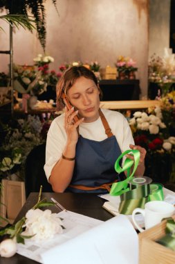 Önlüklü orta yaşlı kadın çiçekçi akıllı telefondan arıyor. Lojistiği ve teslimatı ayarlıyor, emirleri alıyor. Küçük bir yerel işletme. Dizinin bir parçası. Yumuşak odak